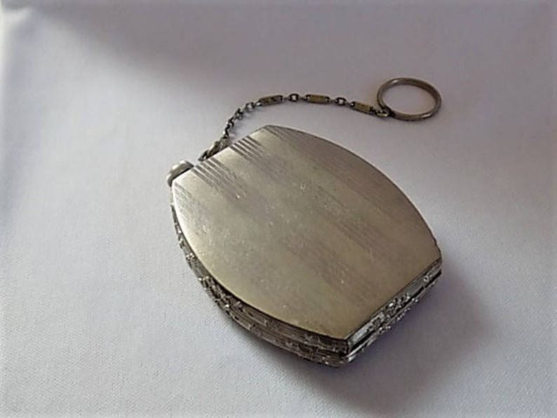 K&ouml;nnte beinhalten: Ein silbernes Kompakt mit einer Kette und einem Ring. Das Kompakt hat eine strukturierte Oberfl&auml;che und ein dekoratives Design an den R&auml;ndern.