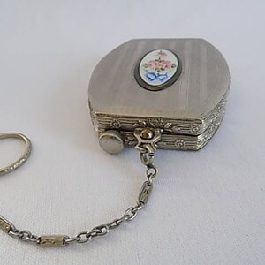 K&ouml;nnte beinhalten: Eine silberne Kompaktdose mit einem Blumendesign auf dem Deckel, die an einer Kette mit einem Ring befestigt ist.