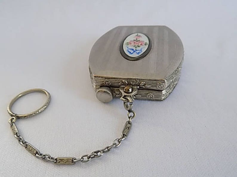 K&ouml;nnte beinhalten: Eine silberne Kompaktdose mit einem Blumendesign auf dem Deckel, die an einer Kette mit einem Ring befestigt ist.