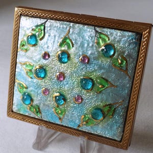 May include: A vintage compact with a gold-toned frame and a blue and green enamel design. The enamel features a floral pattern with blue and green gemstones.
