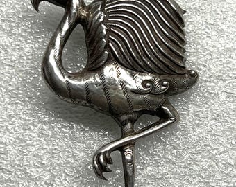 May include: A detailed silver flamingo brooch. The bird is depicted in a standing pose with one leg raised. The brooch has intricate detailing on the feathers and body, with a textured finish. The flamingo's beak is slightly open.