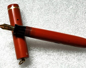 May include: A red fountain pen with a black section and a gold-colored nib. The pen is capped and has a gold-colored ring around the cap.