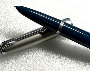 May include: A close-up of a vintage fountain pen with a dark blue barrel and a silver-colored cap. The pen's nib is visible, and the cap is partially open, revealing the pen's inner workings. The pen is a classic writing instrument.