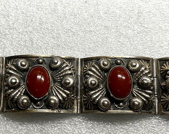 May include: A silver-toned bracelet featuring four rectangular panels, each adorned with a central oval, reddish-brown stone. The panels are intricately detailed with floral and spiral patterns.