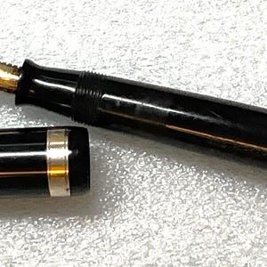 May include: A black fountain pen with a gold nib and accents, disassembled. The pen's cap and body are black, with gold and silver trim. The nib is gold-colored, and the ink feed is yellow.