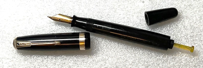 May include: A black fountain pen with a gold nib and accents, disassembled. The pen's cap and body are black, with gold and silver trim. The nib is gold-colored, and the ink feed is yellow.