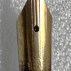 May include: Close-up of a gold-colored fountain pen nib. The nib has a distinctive shape with a small circular breather hole and etched markings. The pen's base is black, and the background is a textured, silvery-white.