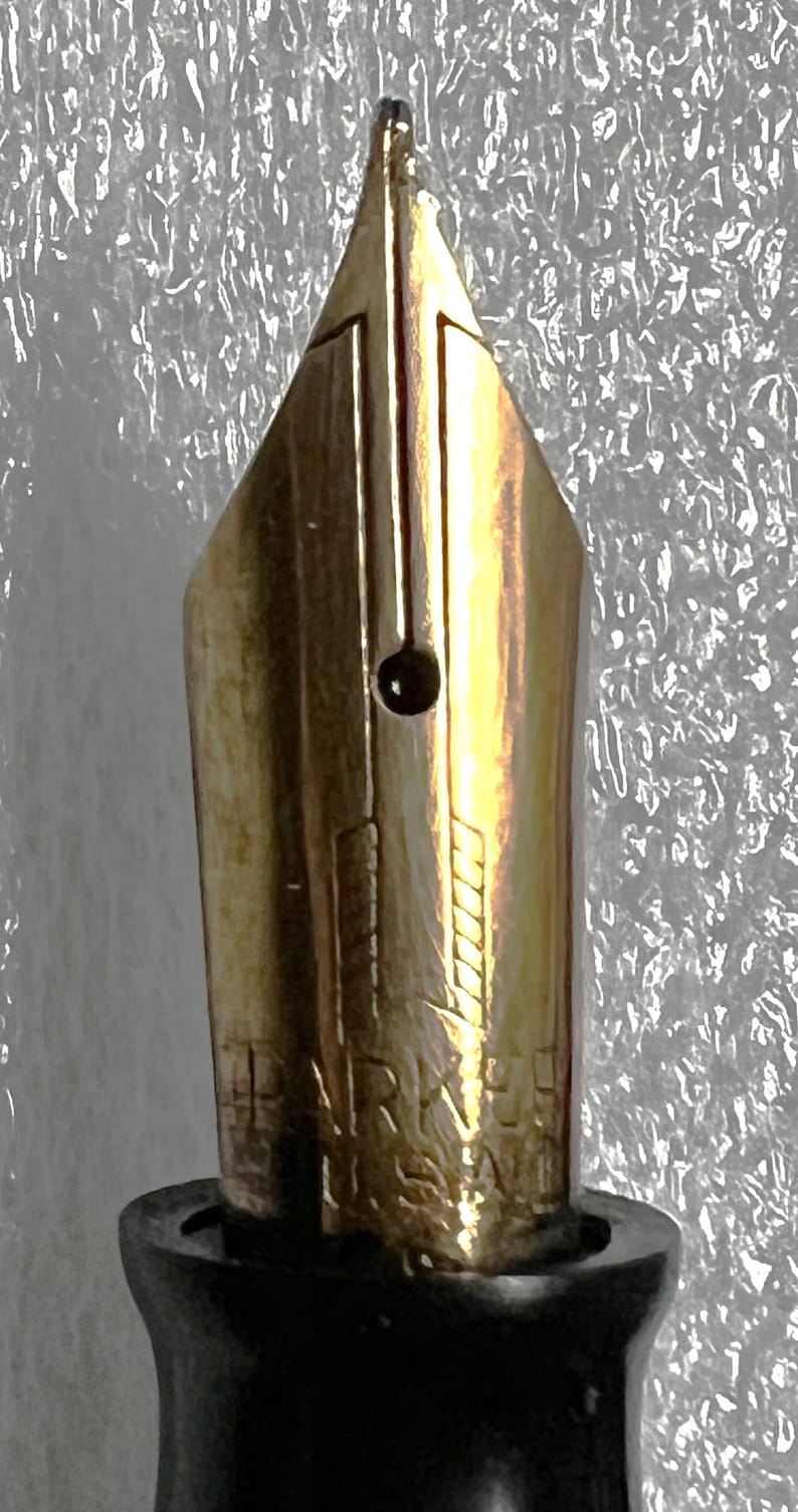 May include: Close-up of a gold-colored fountain pen nib. The nib has a distinctive shape with a small circular breather hole and etched markings. The pen's base is black, and the background is a textured, silvery-white.