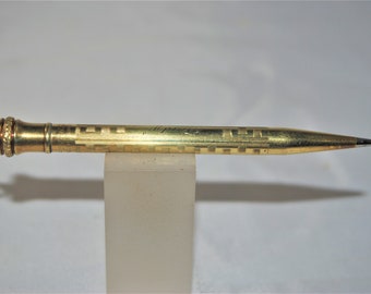 May include: A gold-colored mechanical pencil with a decorative geometric pattern. The pencil has a ring at the end and a pointed tip. The pencil is resting on a clear, rectangular stand.