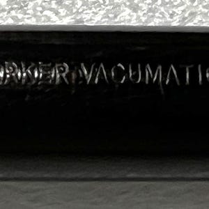 May include: Close-up of a black Parker Vacumatic pen. The pen has the words "PARKER VACUMATIC" and "MADE IN U.S.A." engraved on its surface. The pen's design is sleek and classic, with a focus on the brand's name.