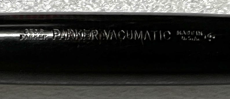 May include: Close-up of a black Parker Vacumatic pen. The pen has the words "PARKER VACUMATIC" and "MADE IN U.S.A." engraved on its surface. The pen's design is sleek and classic, with a focus on the brand's name.