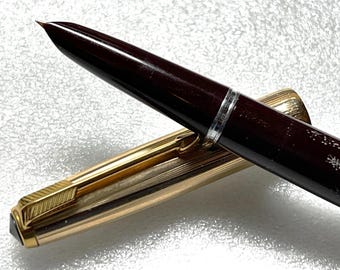 May include: A burgundy fountain pen with gold accents and a silver band. The pen is capped and lying on a white surface.