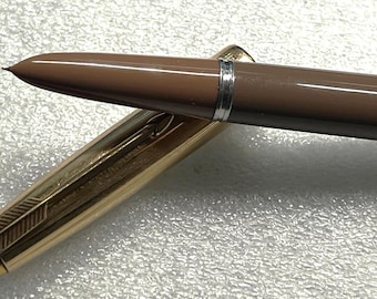 May include: A brown fountain pen with a gold-colored cap. The pen's body is a solid brown color, and the cap has a gold finish with a decorative arrow design. The pen's nib is visible, and the pen is lying on a textured surface.