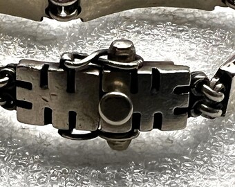 May include: Close-up of a silver bracelet with a unique clasp. The bracelet features a series of rectangular links and a detailed clasp mechanism. The metal has a polished finish, reflecting light. The design is intricate and appears to be handmade.