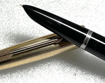 May include: A black fountain pen with a gold-colored barrel and cap. The pen has a silver band near the nib and a gold arrow design on the cap. The pen is lying on a textured surface.