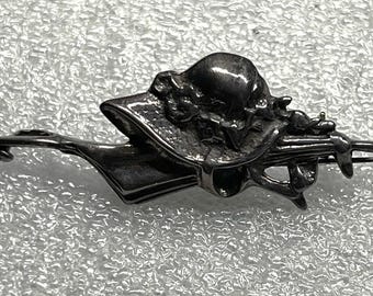 May include: A silver-toned brooch featuring a hat with floral accents and a decorative element. The brooch has a pin closure on the back. The design is detailed and vintage-inspired.
