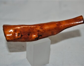 May include: A handcrafted, wooden pipe with a polished, glossy finish. The pipe is a warm, medium brown color with darker knots and variations in the wood grain. It is displayed on a small, white, rectangular stand.