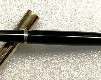 May include: A black fountain pen with a gold-colored cap. The pen is lying on a white, textured surface. The pen's body is black with a silver band near the nib.