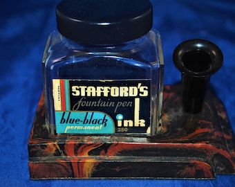 May include: A vintage inkwell set with a glass ink bottle and a black pen holder. The ink bottle is filled with blue-black ink and has a label that reads "Stafford's fountain pen blue-black permanent ink 250". The inkwell set is made of black and red marbled plastic.