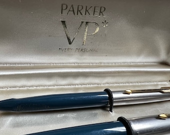 May include: Two Parker VP pens in a white satin lined box. The pens are silver with blue barrels and gold accents. The box lid has the Parker logo and the text "VP* Very Personal".