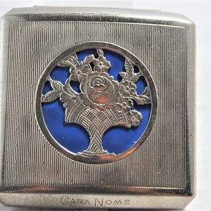 May include: Silver compact with a blue enamel floral design and the words "Cara Nome" engraved on the bottom.