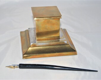 May include: A brass inkwell with a hinged lid and a square base. A black ink pen is lying on a white surface next to the inkwell.