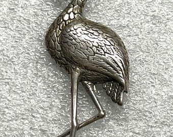 May include: A silver-toned flamingo brooch. The bird is depicted in a standing pose with its head curved back. The brooch has detailed feather and body textures. The flamingo's legs are thin and delicate.