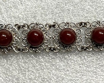 May include: A silver-toned bracelet featuring seven round, deep red cabochon stones set within ornate, openwork rectangular links. The bracelet has a secure clasp closure. The design is vintage-inspired and elegant.