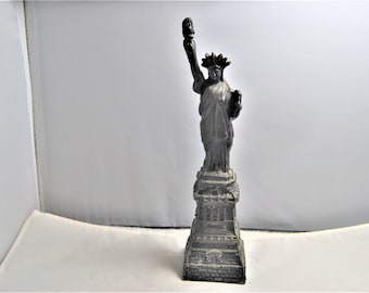 May include: A small, gray metal statue of the Statue of Liberty. The statue is holding a torch in her right hand and a book in her left hand. The statue is standing on a base with a detailed design.