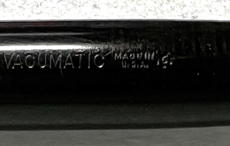 May include: Close-up of a black pen with the word "VACUMATIC" engraved on the side. The text "MADE IN U.S.A." is also visible. The pen has a shiny, reflective surface.