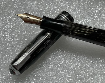May include: A black and silver fountain pen with a gold nib and a striped pattern. The pen is lying on a white textured surface.