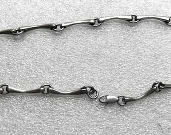 May include: A silver-toned necklace with a unique design. The necklace features a series of curved, elongated links connected by small circular rings. The clasp is a simple, secure design. The necklace is displayed on a textured, light-colored surface.