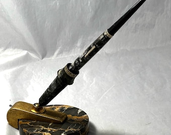 May include: An antique fountain pen with a black and gold marbled design, resting on a matching pen stand. The pen features a black nib and gold accents. The pen stand has a gold-colored pen rest and a marble base.
