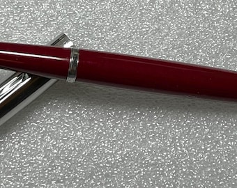 May include: A red fountain pen with a silver cap and barrel. The pen is on a white textured surface.