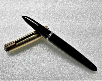 May include: A black and gold fountain pen with a cap on the barrel. The pen is on a white textured surface.
