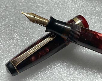 May include: Close-up of a vintage fountain pen with a gold nib and a maroon and black marbled barrel. The pen has a gold clip and a black section where the nib is held. The pen is a classic writing instrument.