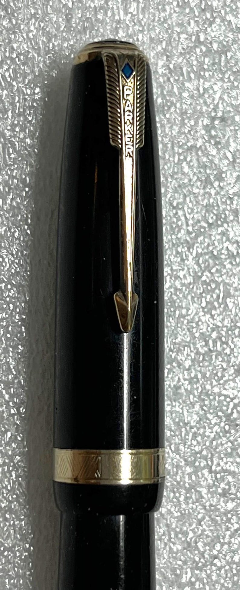 May include: Close-up of a black Parker pen with gold accents. The pen features a gold arrow clip and a gold band near the base. The top of the pen has the Parker logo and a blue accent.
