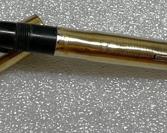 May include: A gold-colored fountain pen with a black grip and gold nib. The pen's body has a textured, ribbed design. The cap is open, revealing the nib. The pen is lying on a textured surface.