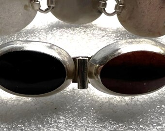 May include: A silver bracelet featuring oval links with black and brown insets. The bracelet is composed of multiple oval-shaped links connected by smaller circular links. The clasp is a simple bar and loop design.