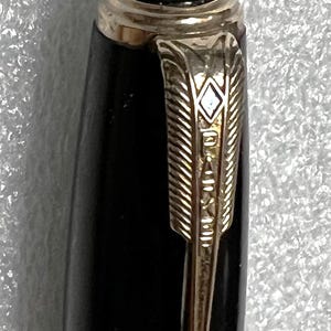 May include: Close-up of a black pen with gold-colored accents. The pen features a gold-colored arrow-shaped clip with the brand name engraved. The top of the pen has a black cap with a gold band.
