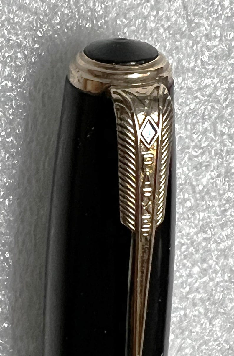 May include: Close-up of a black pen with gold-colored accents. The pen features a gold-colored arrow-shaped clip with the brand name engraved. The top of the pen has a black cap with a gold band.