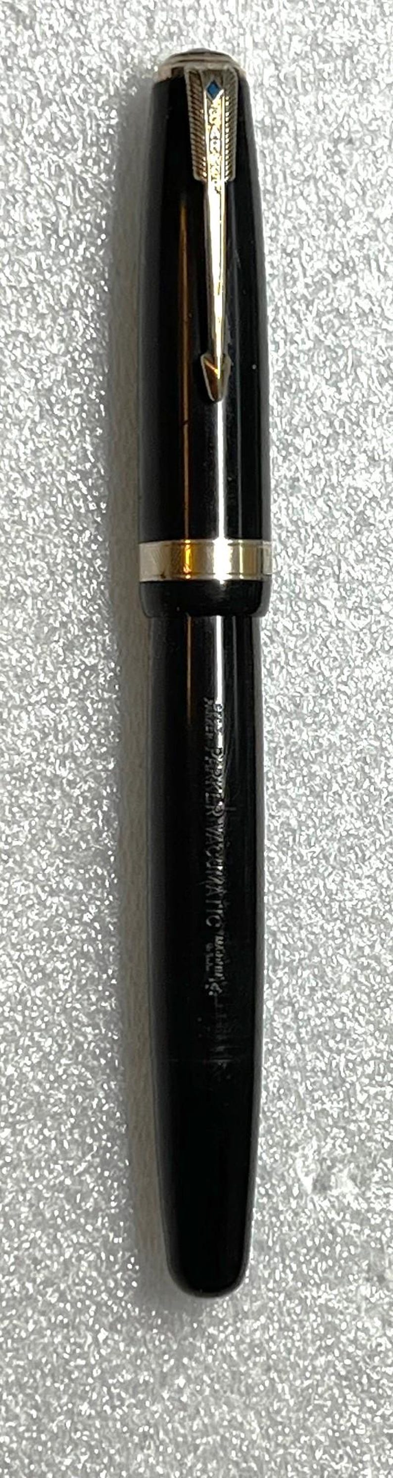 May include: A black fountain pen with gold-colored accents. The pen has a gold clip and a gold band near the center. The pen's nib is visible, and the pen's body has the text "PARKER VACUMATIC" printed on it.