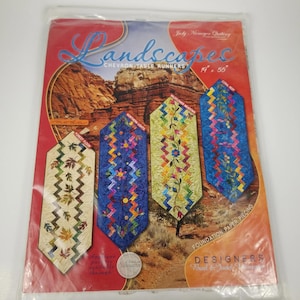 May include: A package featuring four chevron table runner designs. The designs include "Forest Floor," "Northern Lights," "Wildflower Meadows," and "Coral Reef." The package is labeled "Landscapes" and measures 19 x 55 inches.