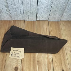 May include: A dark brown, elongated, open-top fabric container with a structured, angled silhouette. A small, square, off-white paper tag is attached, featuring the printed text "Yarn-Craft Accessories, Support Your Independent Yarn Store, YARN-CRAFT SUPPLIES, Milwaukee, Wisconsin."