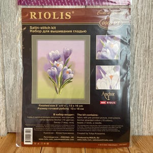 May include: A Riolis satin-stitch kit in a clear plastic package featuring a floral design of purple crocus flowers. The packaging displays the finished size of 5 inches by 6.25 inches, 13 cm by 16 cm, and includes Anchor stranded cotton threads.