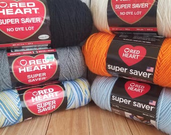 May include: A collection of Red Heart Super Saver yarn skeins in various colors, including black, white, gray, orange, blue, and a multi-colored skein. The labels indicate the yarn is worsted weight and no-dye-lot.