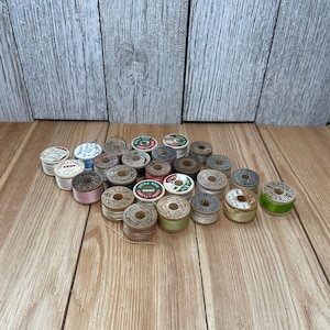 May include: A collection of vintage wooden and cardboard spools of sewing thread in various colors including white, pink, green, and tan. Each spool features a circular paper label on top with printed brand names and text details.