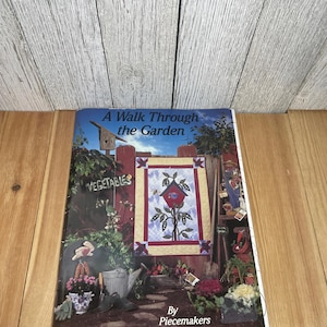 May include: A softcover book titled A Walk Through the Garden by Piecemakers. The cover features a quilt with a birdhouse and plant motif, set against a garden background with a wooden fence, a sign reading Vegetables, and various plants.
