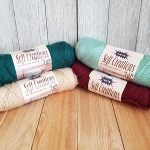 May include: Four skeins of Soft Creations yarn in various colors: teal, light green, cream, and burgundy. Each skein has a label with the brand name and text. The yarn is arranged on a wooden surface with a white wood background.