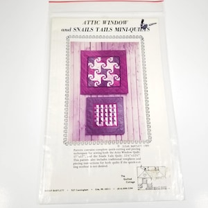 May include: A clear plastic bag containing a pattern for mini-quilts. The pattern includes instructions for "Attic Window" and "Snails Tails" quilts. The pattern features two quilt designs in shades of purple and white. Text on the pattern reads "The Quilted Cottage."